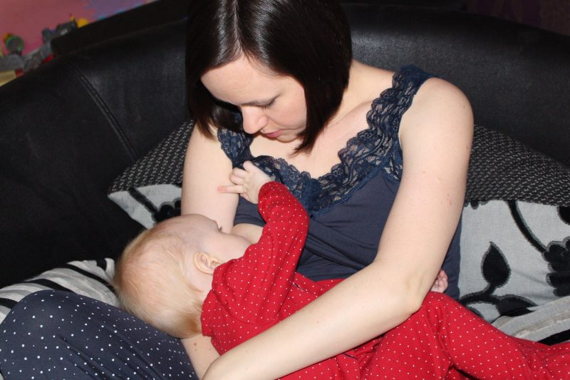 A brunette woman nursing her baby who is in a red sleep suit
