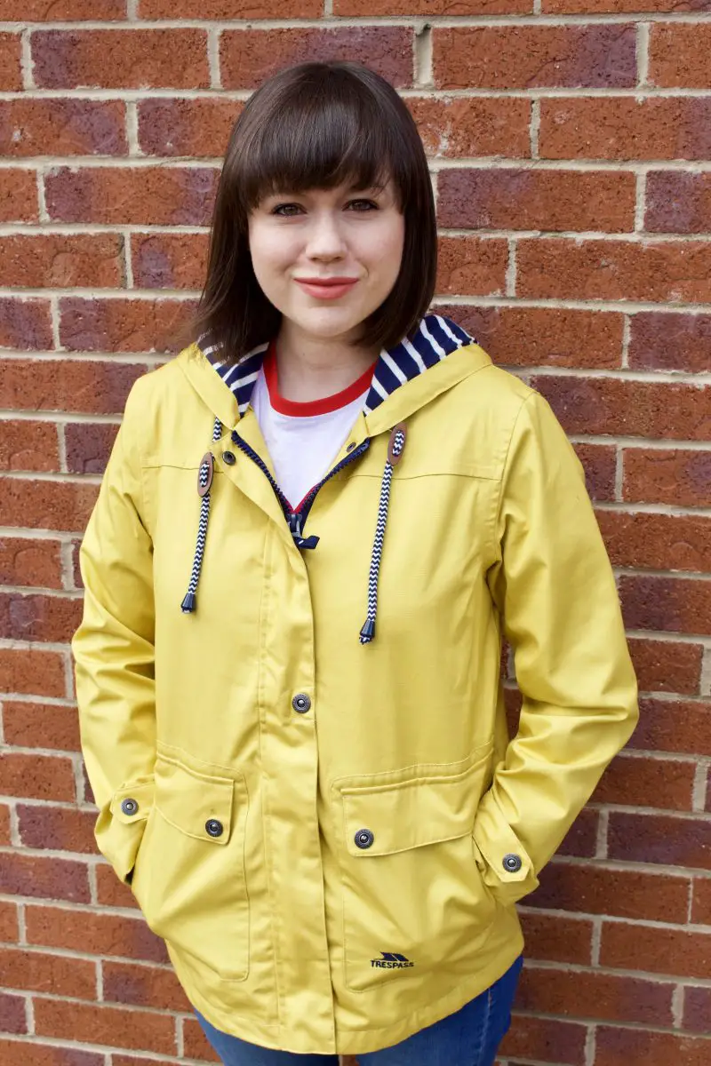 A brunette woman smiles at the camera wearing a yellow Tresspass Seawater jacket. You can see blue and white striped toggles and inner hood lining. 