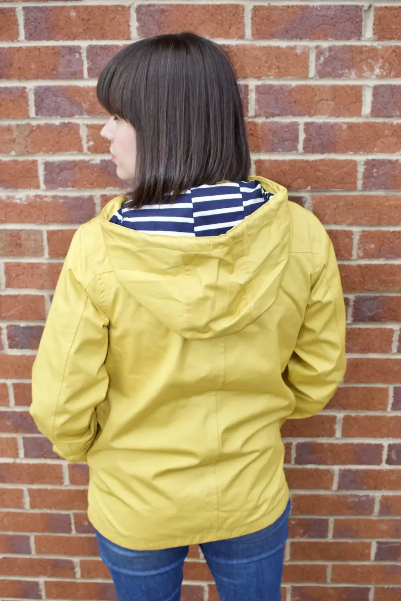 A brunette woman has her back to the camera, she is wearing a yellow Tresspass Seawater jacket with a blue and white striped inner hood. 