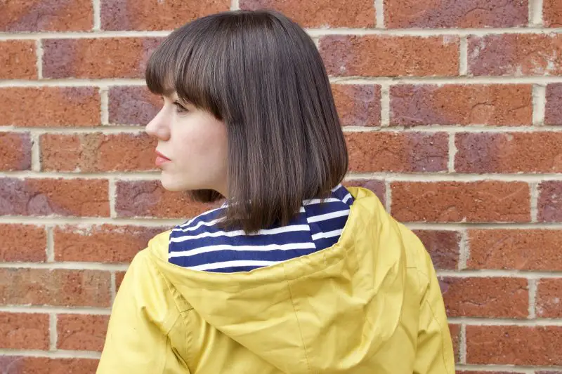 A brunette woman has her back to the camera, she is wearing a yellow Tresspass Seawater jacket with a blue and white striped inner hood. 