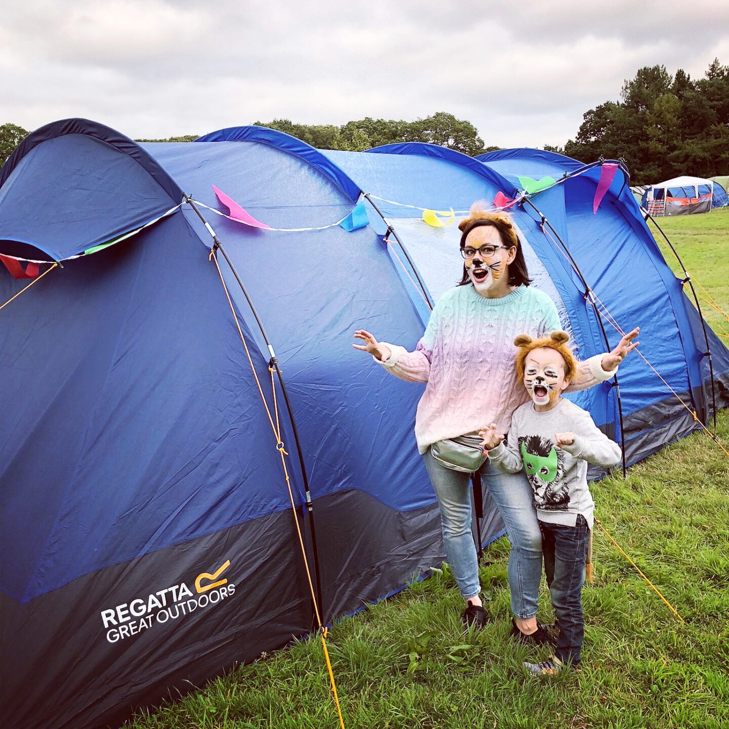 Woman and child wearing lion face faints, pretending to roar whilst stood in front of a blue tent with rainbow coloured bunting. 