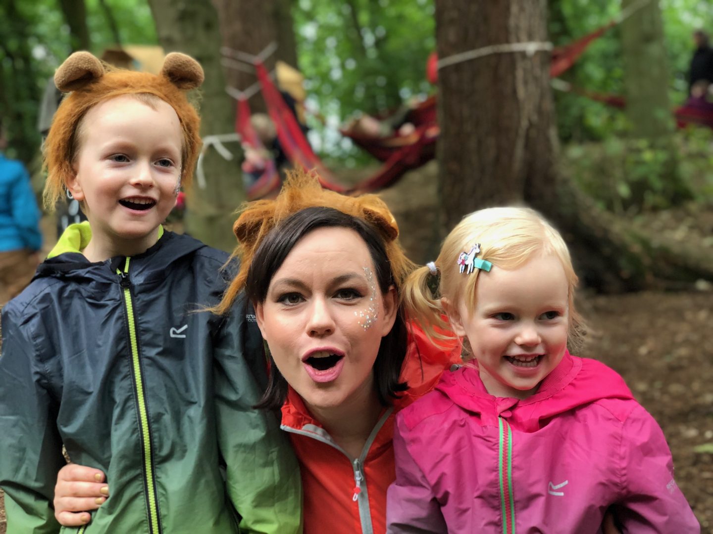 A woman and a boy and a girl and all posing for a photo together and are laughing. They are clearly at a festival and are wearing waterproof jackets. 