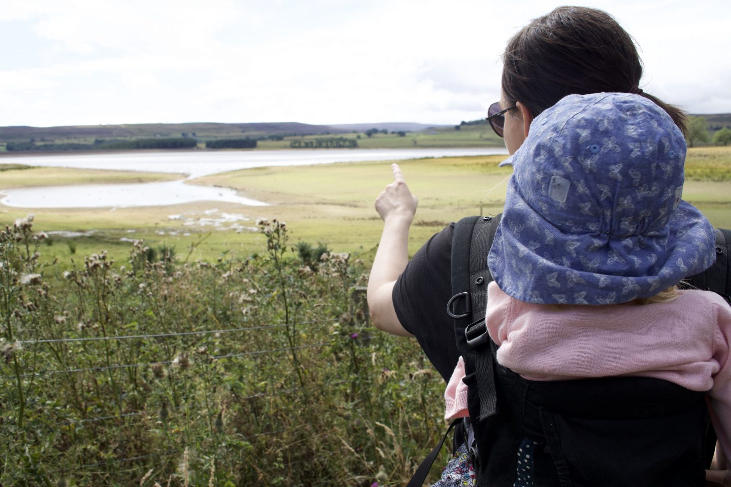 A woman carrying a toddler in a baby carrier on her back whilst pointing at the reservoir 