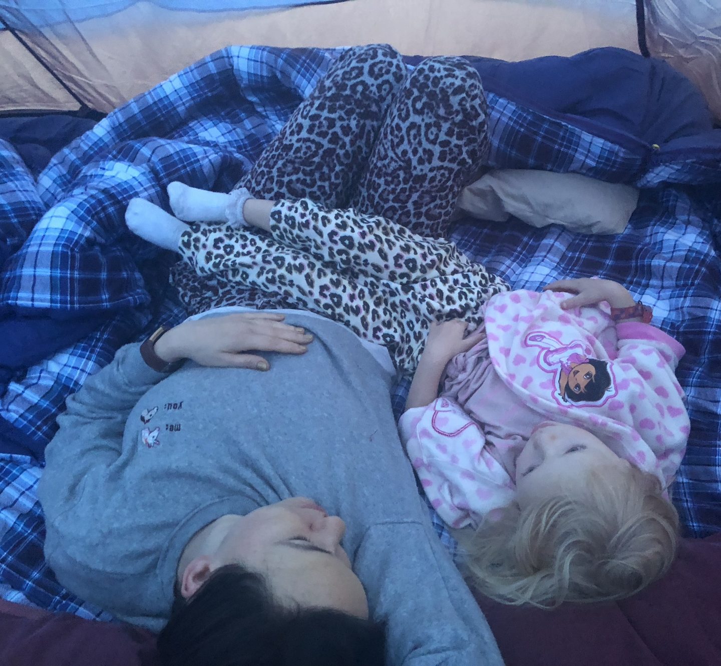 A woman and a girl laid together inside a tent on sleeping bags. They look cosy and the girls legs are draped across her mother's stomach. 