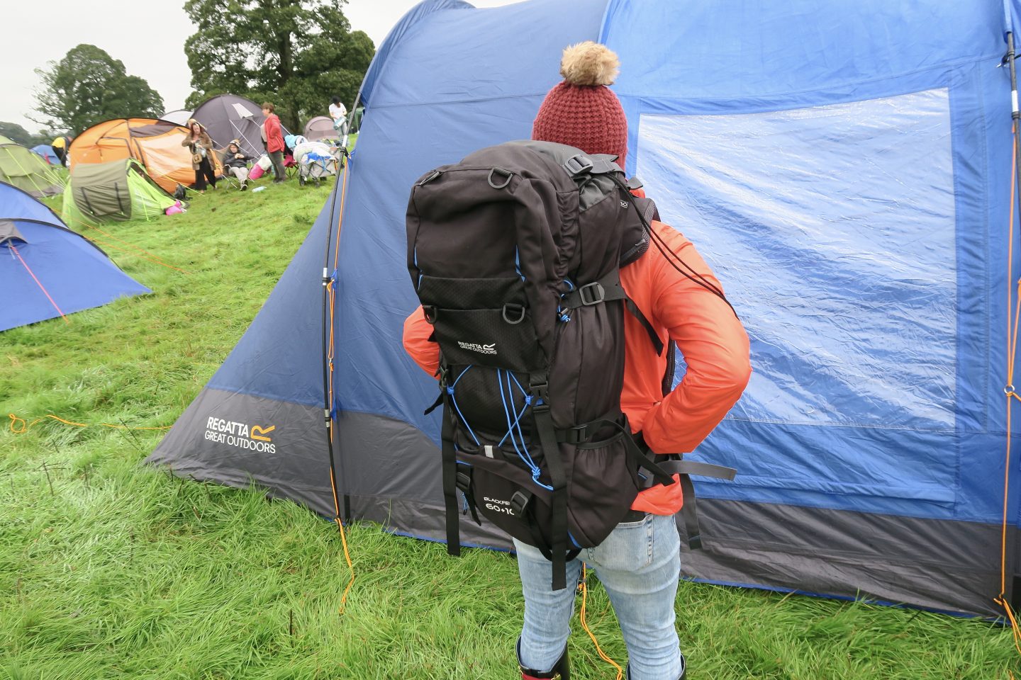 A woman wears a camping rucksack in front of her tent at a festival. 