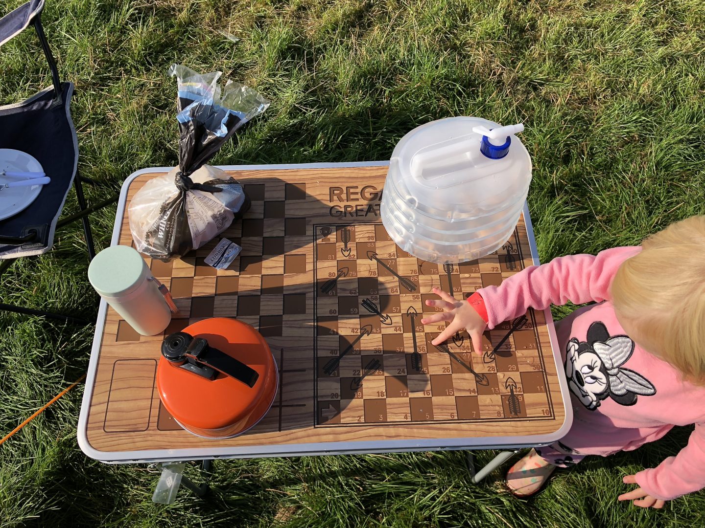 A toddler plays with the checkerboard on a camping games table. The table is on the grass and has a kettle and water canteen on it. 