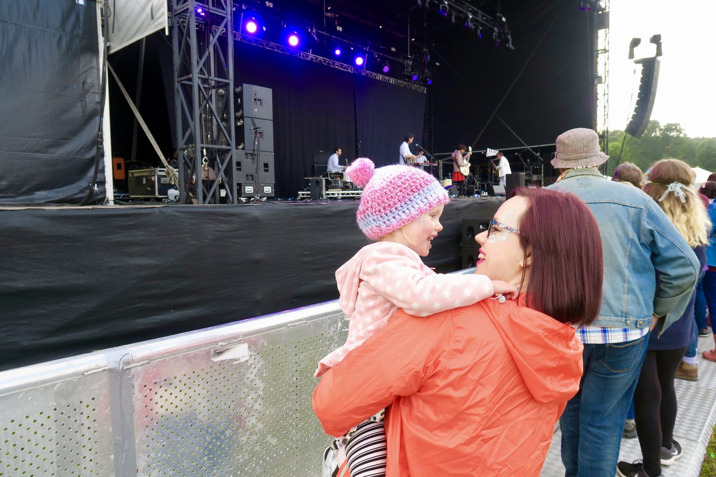 Woman carrying little girl in front of the main stage at a festival. They are both smiling. 