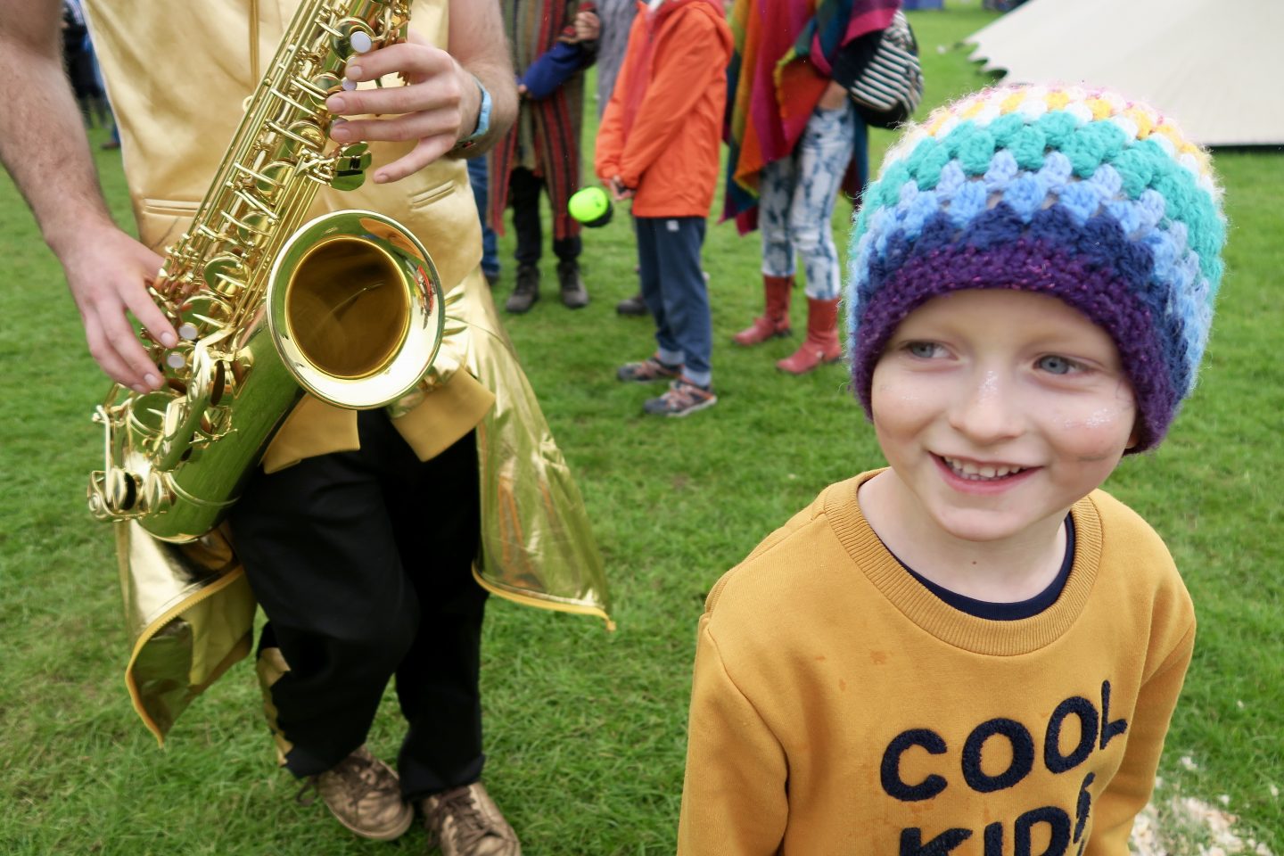 Little boy at a family friendly festival smiling next to a man playing a trumpet. 