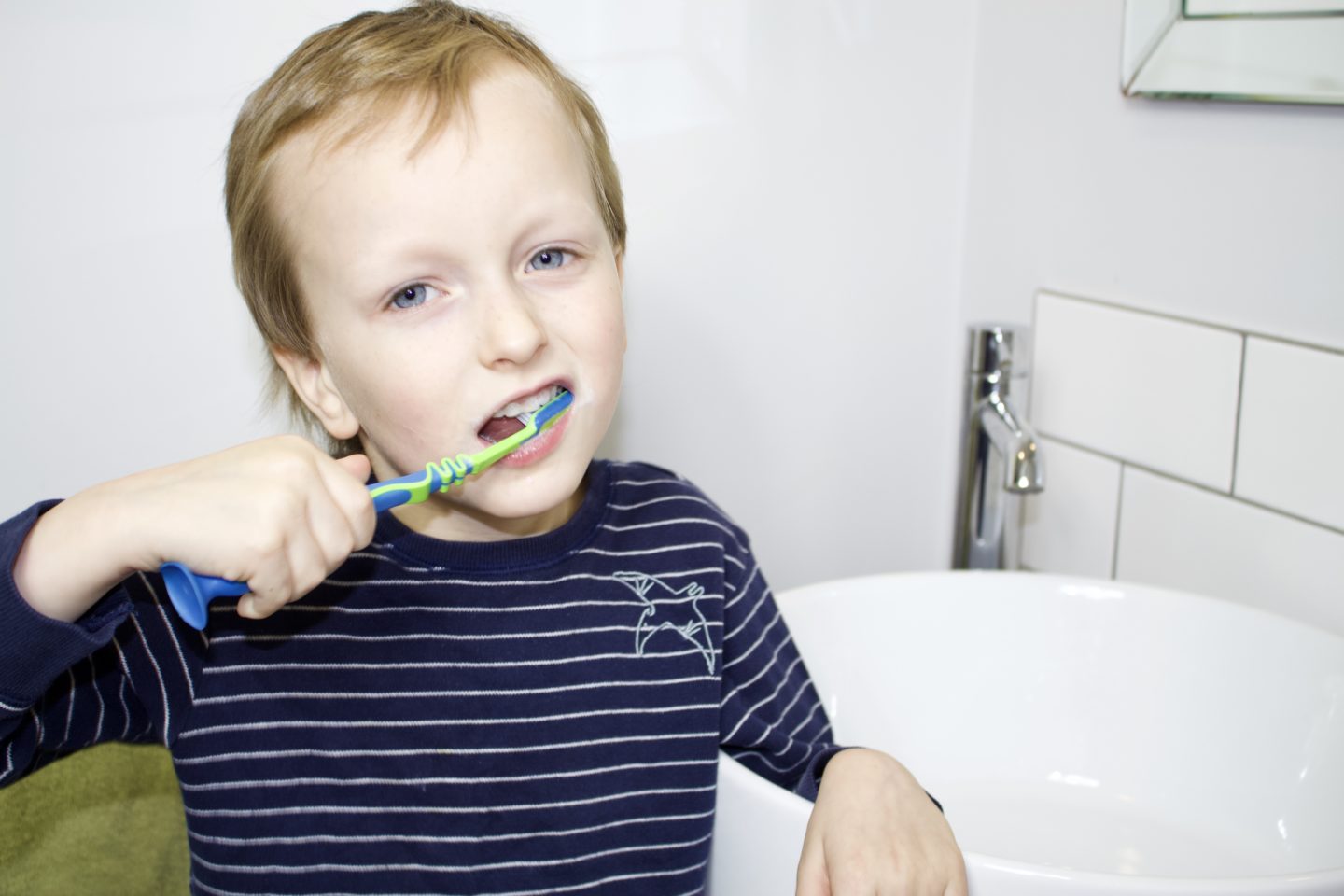 Blonde boy brushing his teeth with a green and blue brush